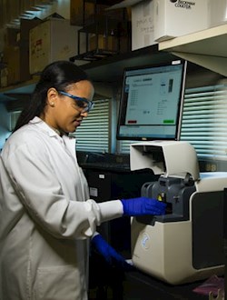 Donna con camice, occhiali e guanti lavora in laboratorio con 2 pc e uno strumento_CTIS, la nuova piattaforma NTT DATA per le sperimentazioni cliniche della European Medicine Agency (EMA)